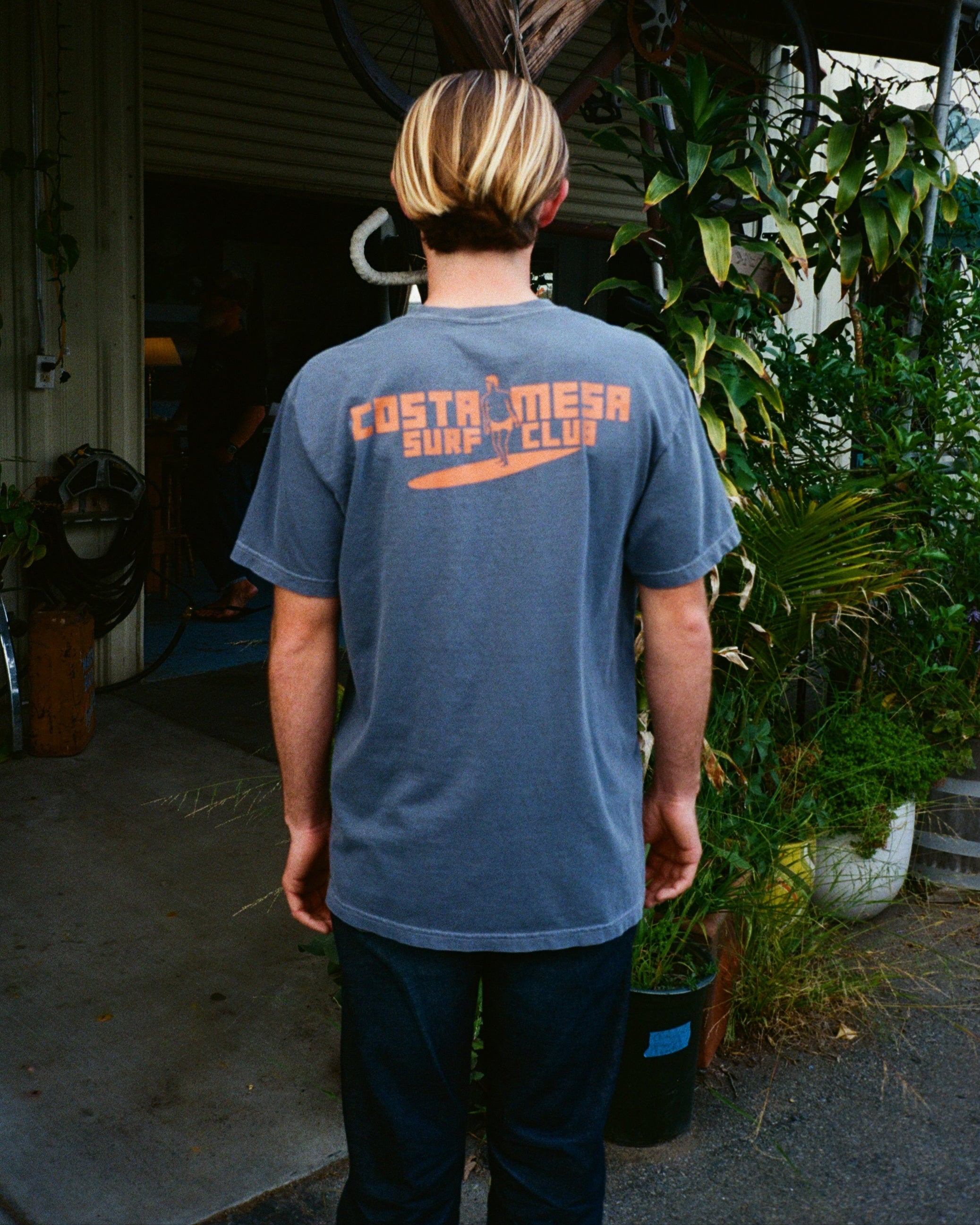 Person wearing a blue t-shirt with orange text, standing outdoors near plants and a wooden structure.