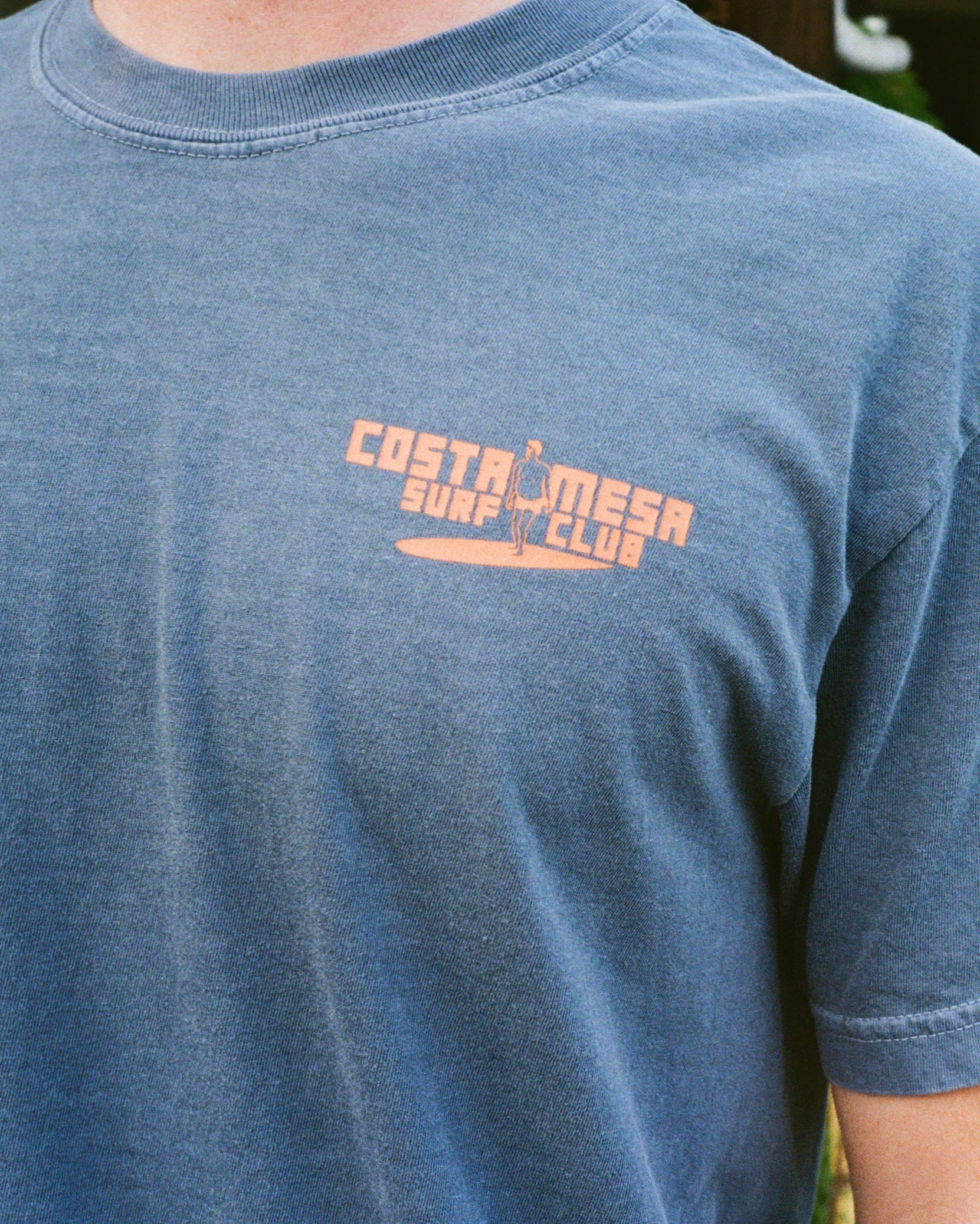 Person wearing a blue t-shirt with 'Costa Mesa Surf Club' logo against a green leafy background