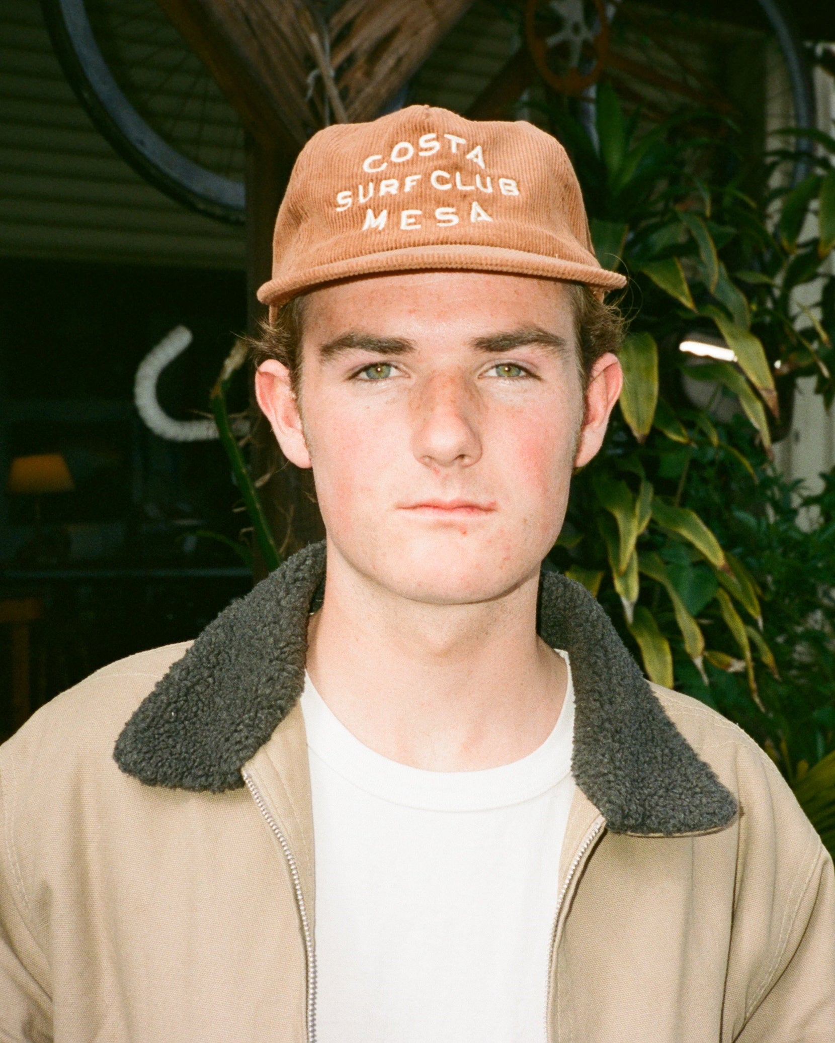 Person wearing a brown cap with 'Costa Mesa Surf Club' text, standing outdoors with greenery in the background.