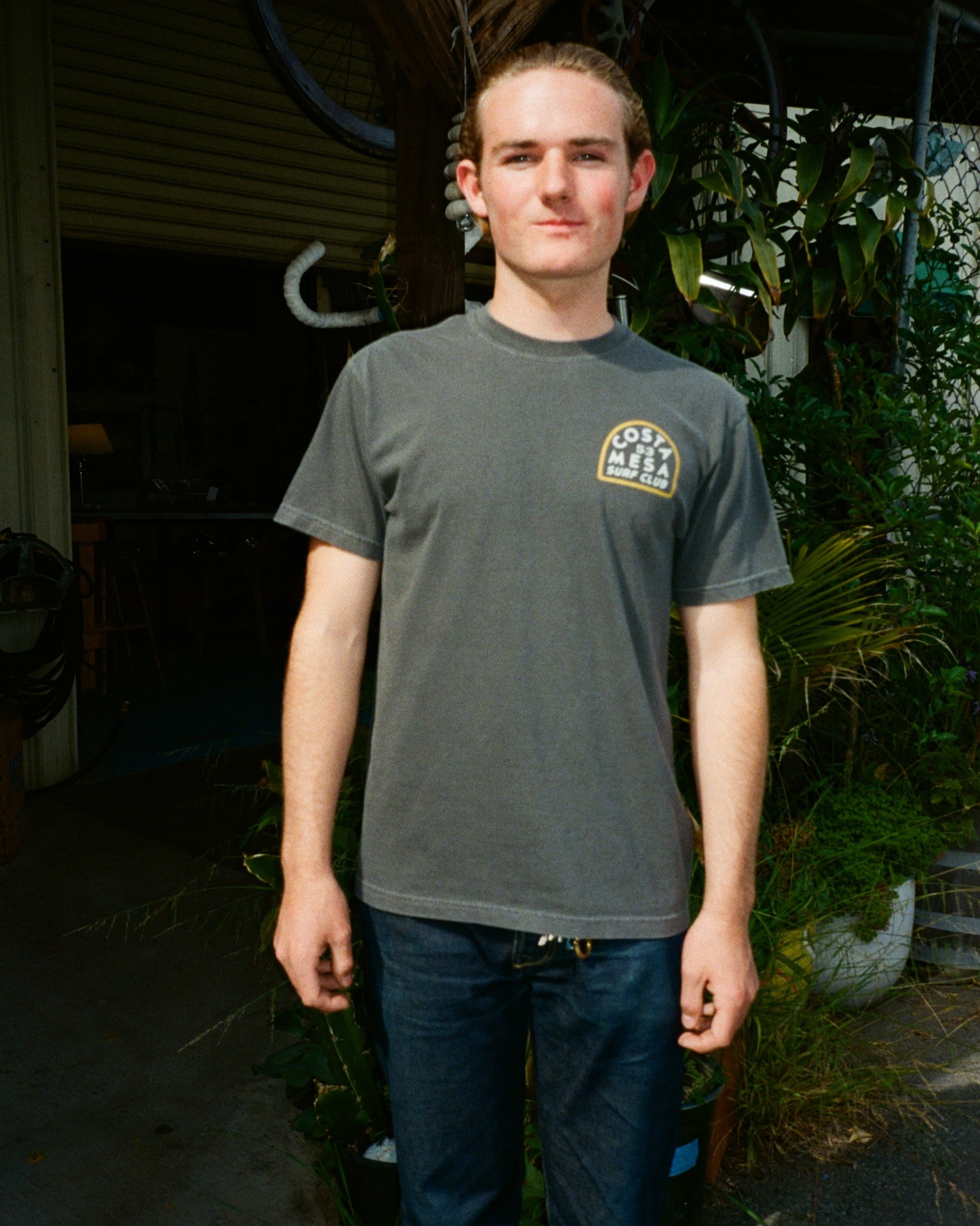 Person wearing a gray t-shirt with a logo, standing outdoors with plants in the background