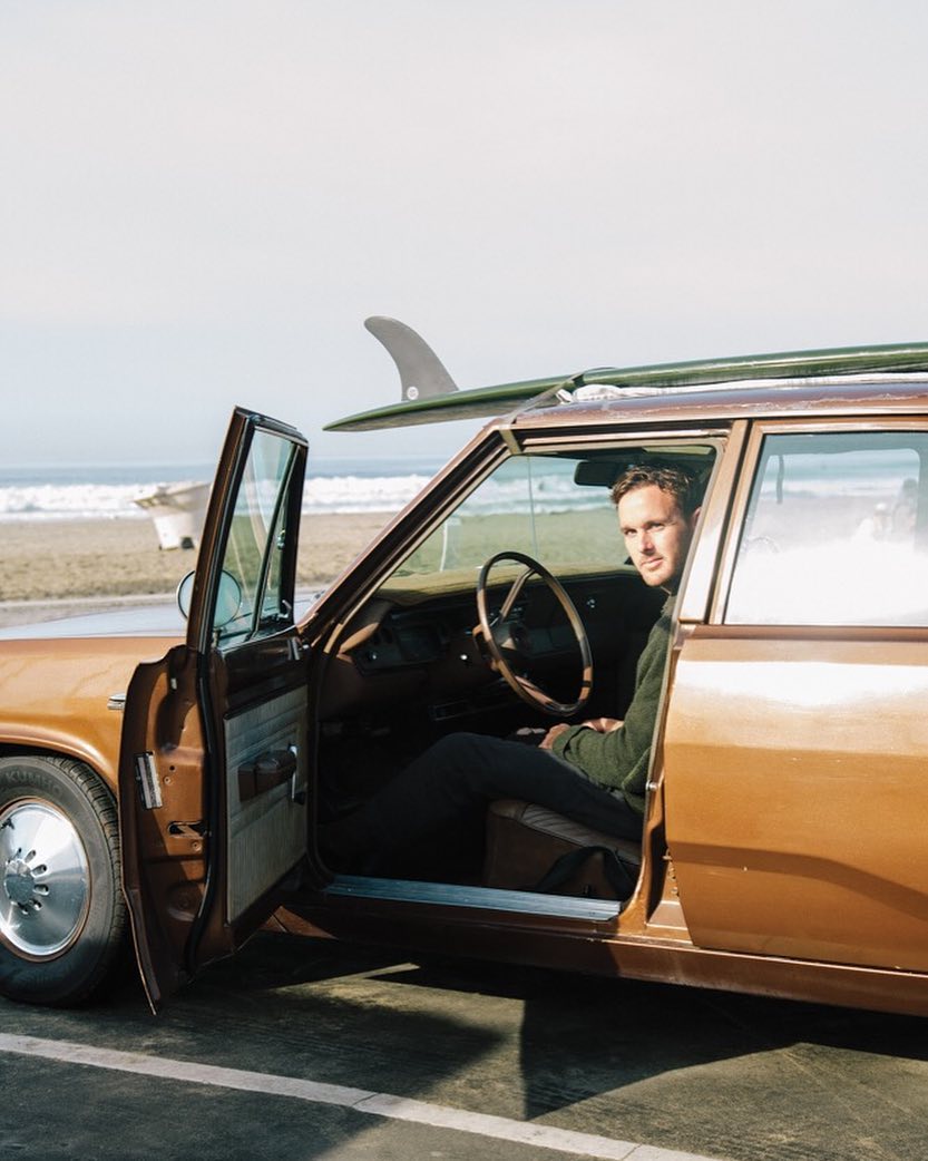 Man sitting in a vintage car with a surfboard on the roof, near a beach.