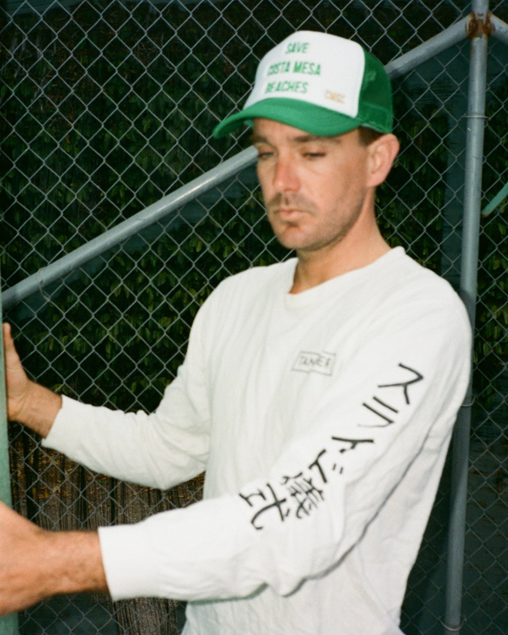 Man wearing a white sweatshirt and green cap with text, standing against a chain-link fence.
