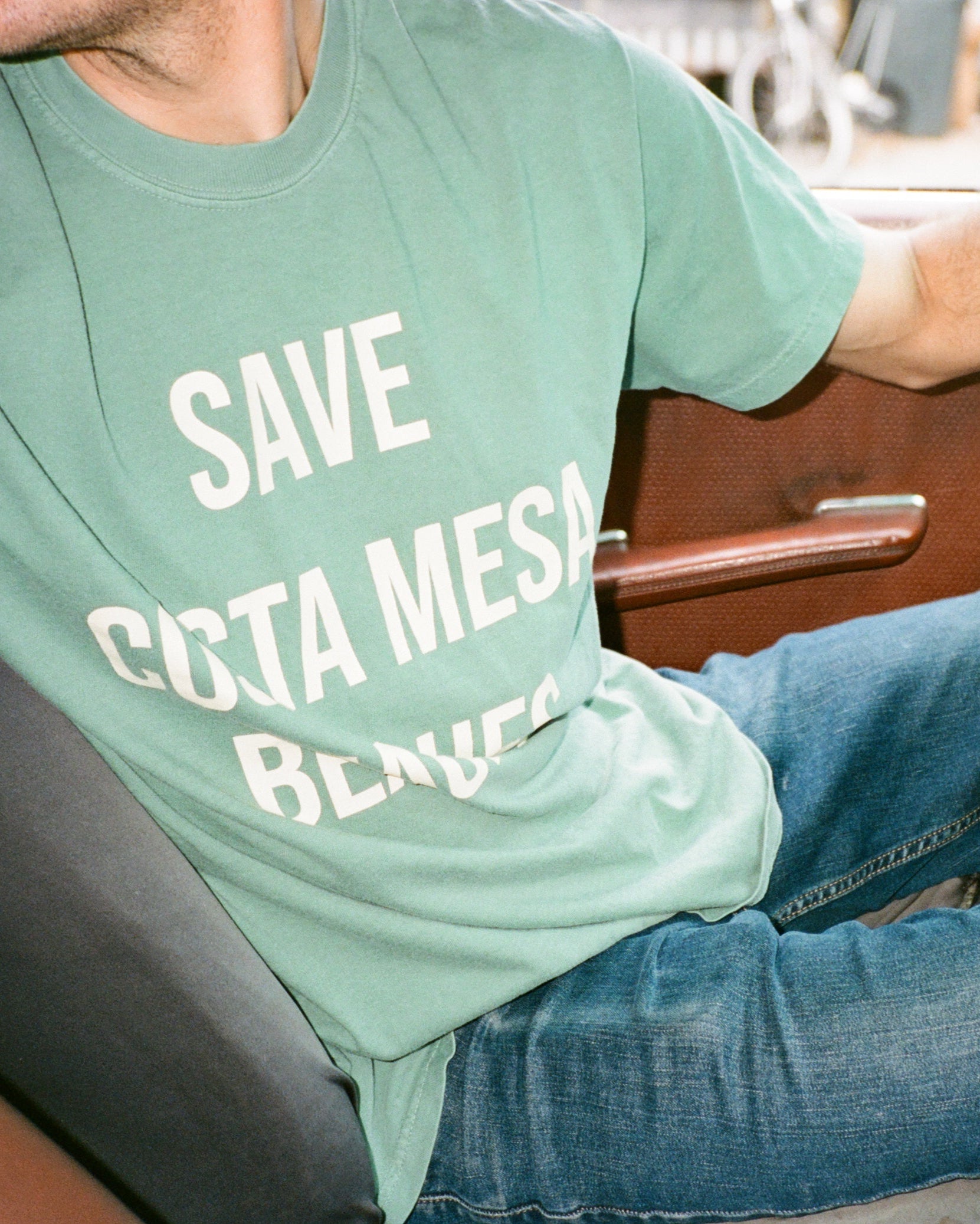 Person sitting in a car wearing a green t-shirt with 'SAVE COSTA MESA BEACHES' text.