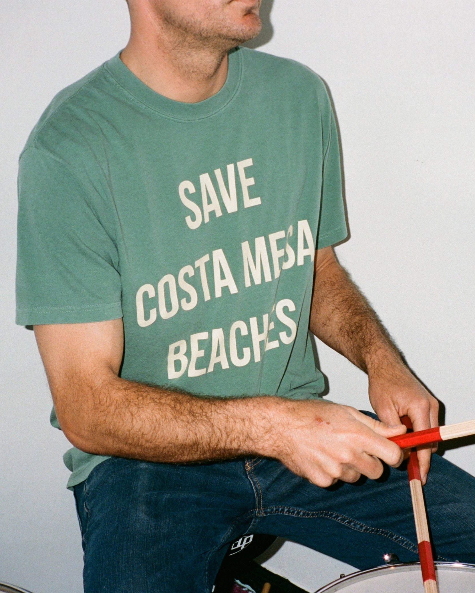 Person playing drums wearing a green t-shirt with 'Save Costa Mesa Beaches' text.