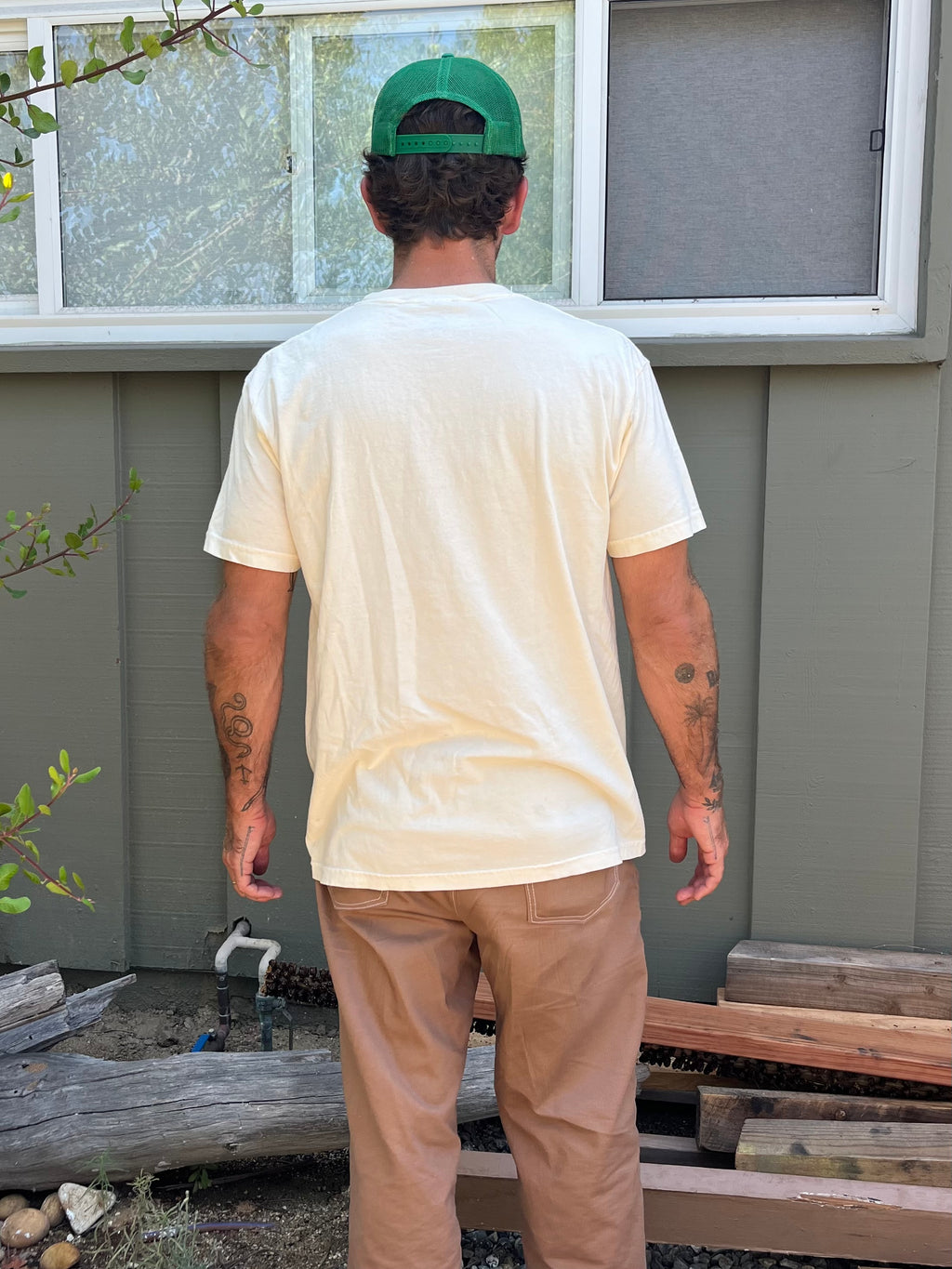 Man wearing a beige t-shirt and brown pants standing in front of a gray wall with a window.