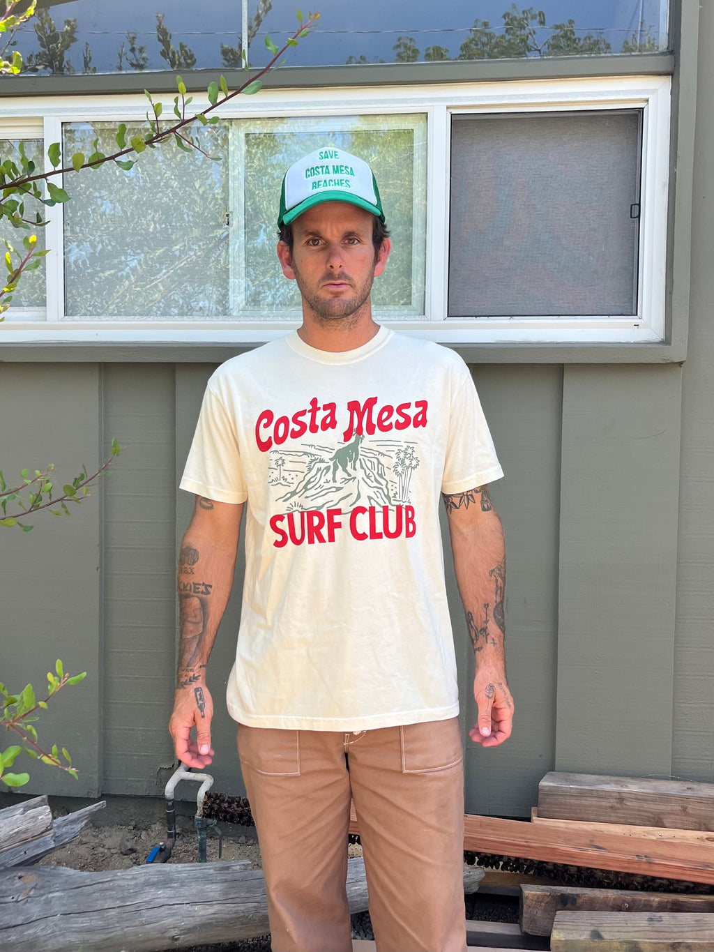 Man wearing a 'Costa Mesa Surf Club' t-shirt standing in front of a house.