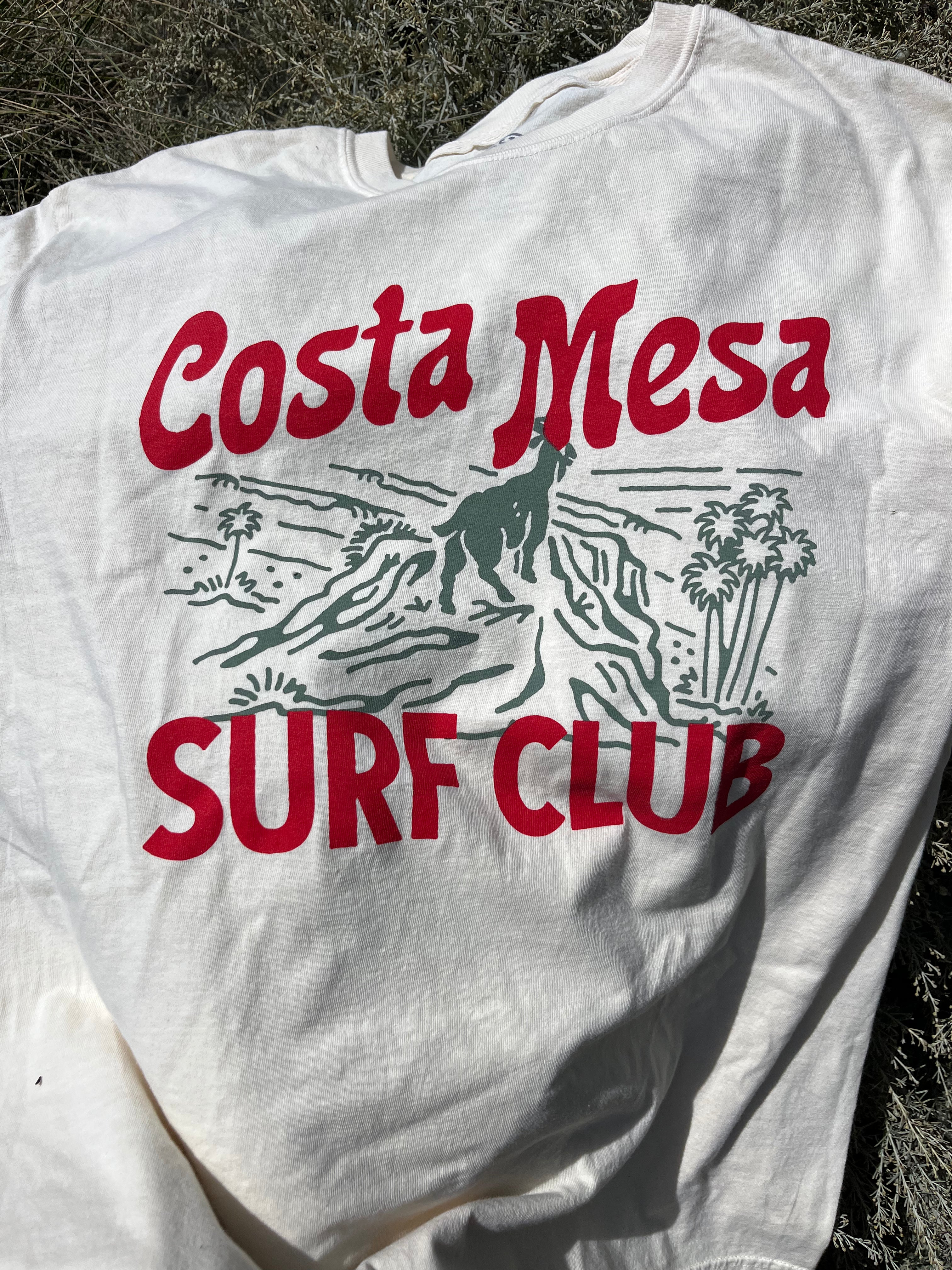 White t-shirt with 'Costa Mesa Surf Club' text and graphics on a natural background